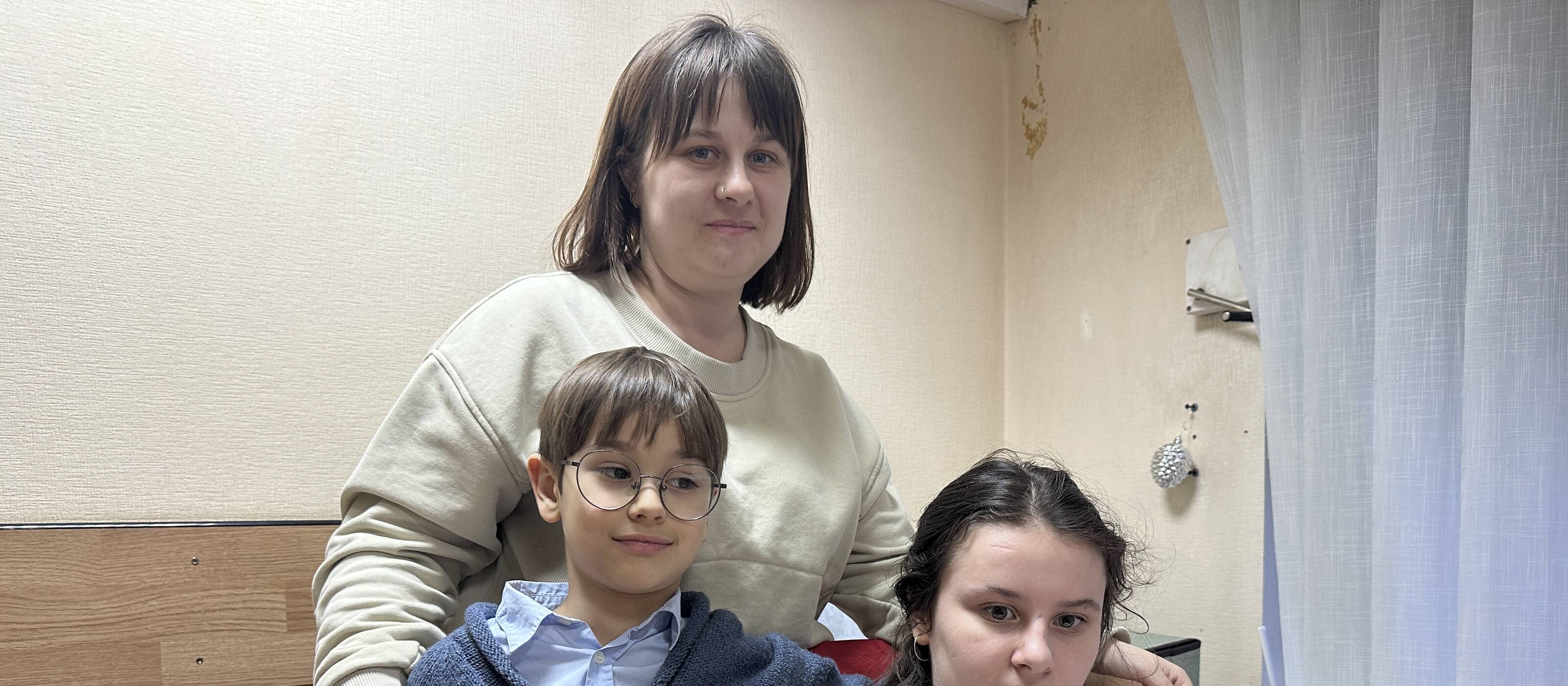 Eine Frau, ein Junge und ein Mädchen schauen in die Kamera. Die Frau und der Junge stehen, das Mädchen sitzt am Tisch und hat eine Zeichnung vor sich.