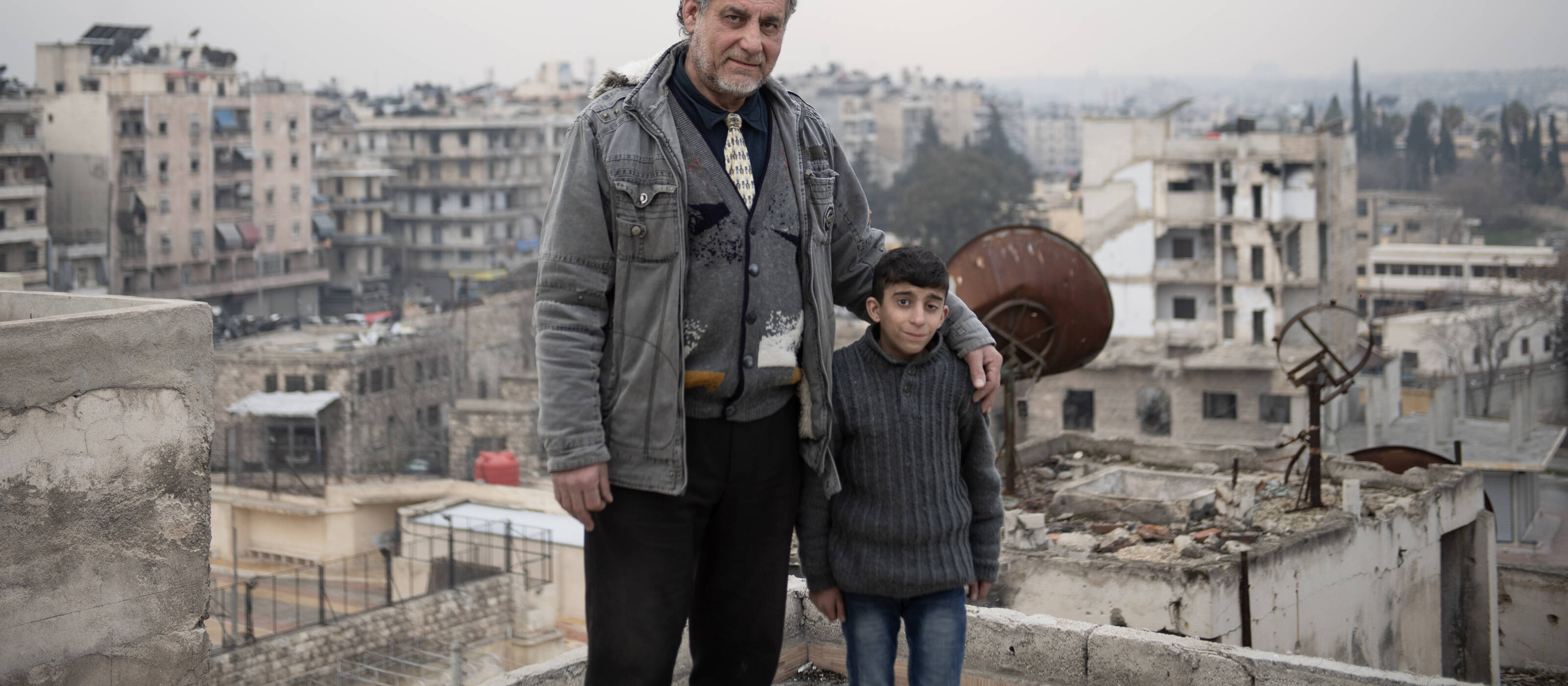 An elderly man looks into the camera, his arm around a boy. The two are standing in front of bombed-out buildings.