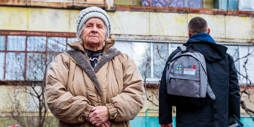 Eine ältere Ukrainer und ein Caritas-Ukraine-Mitarbeiter stehen vor einem zerstörten Gebäude